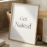 Minimal bathroom wall art featuring the words “Get Naked” in elegant serif typography, framed in light wood and placed on a neutral bathroom floor with soft natural light.