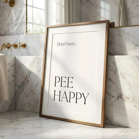 Humorous bathroom wall art print saying “Don’t hurry, Pee Happy,” leaning against a tiled wall near a sink in soft daylight.