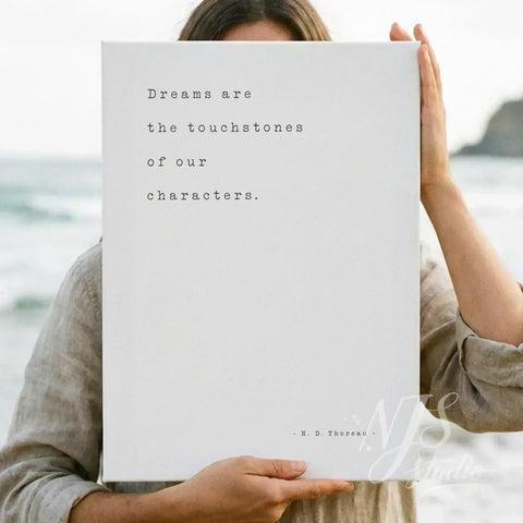 Woman holding a white canvas print with the quote Dreams are the touchstones of our characters on a beach.
