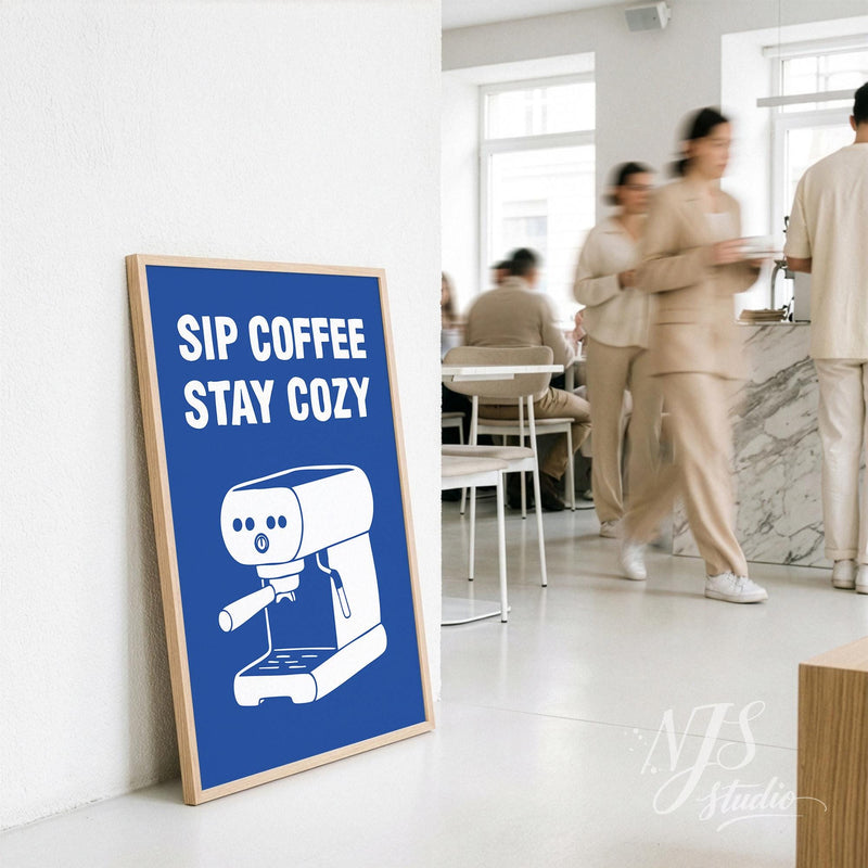 Framed poster with 'Sip Coffee Stay Cozy' text and coffee machine illustration in a modern cafe setting.