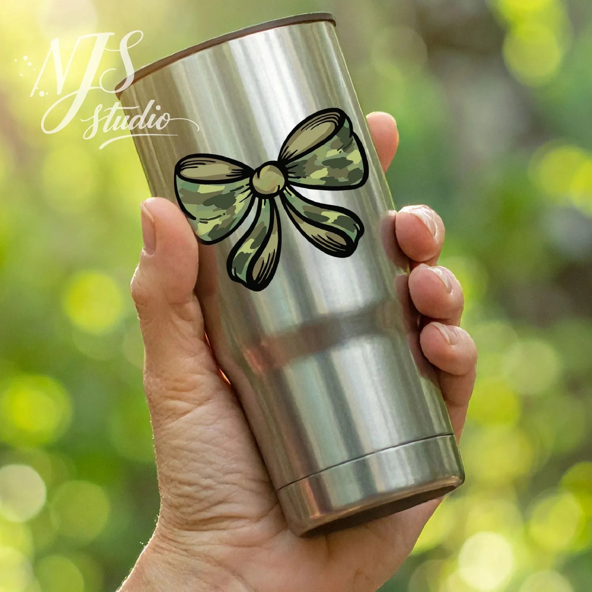 Stainless steel travel tumbler with a camouflage bow design held in hand outdoors with greenery in the background