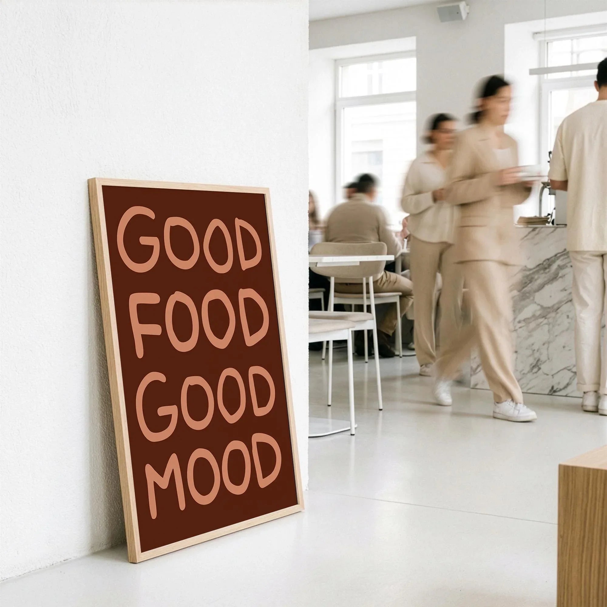 Large wood framed funny kitchen poster standing on the floor in a modern cafe interior with people blurring in background.
