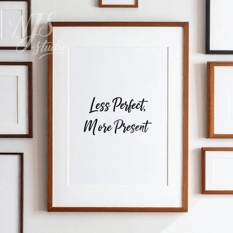 Large framed "Less Perfect More Present" print hanging on a wall surrounded by several smaller art frames, stylish gallery wall decor.