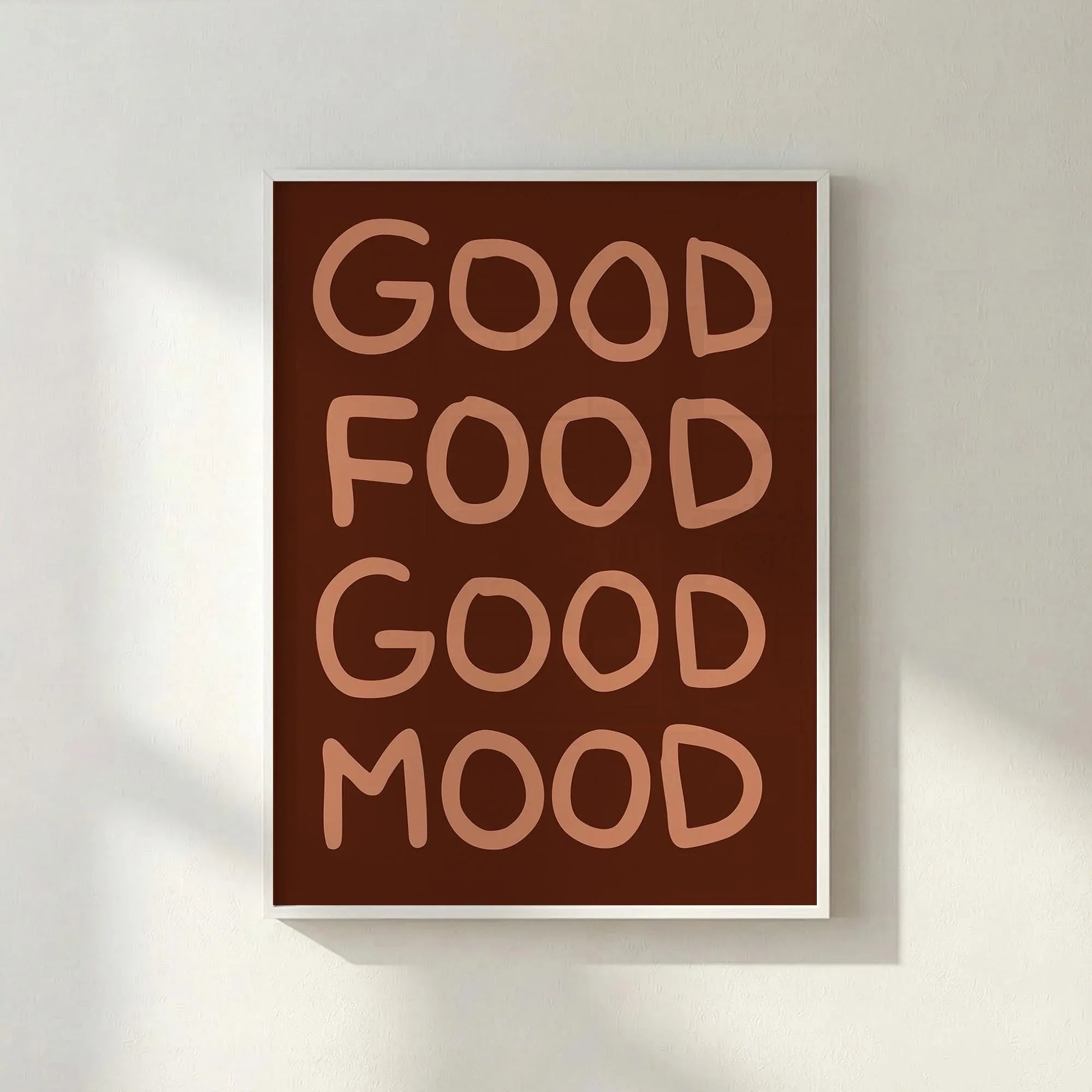 White framed brown typography poster saying Good Food Good Mood leaning against a white textured wall.
