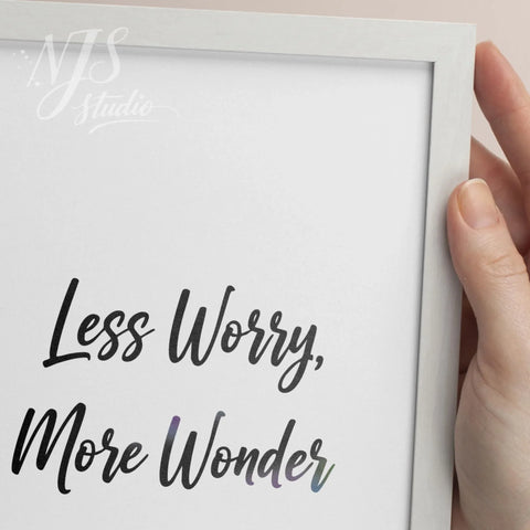 Close up of hands holding a white framed print, showing the hand lettering detail of Less Worry More Wonder.