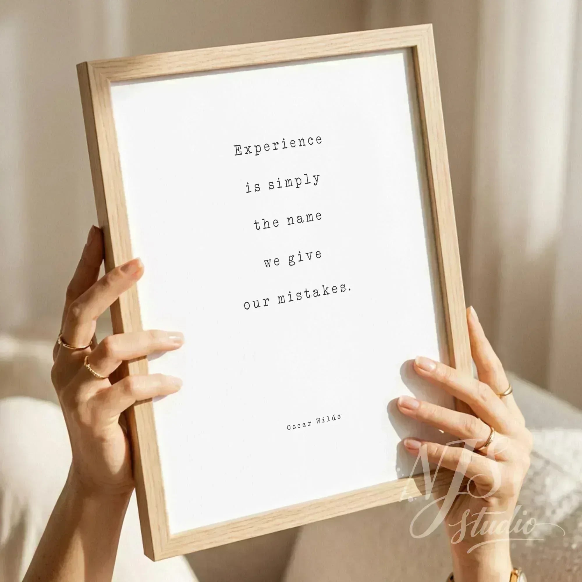 Hands holding a light wood framed print with typewriter text reading Experience is simply the name we give our mistakes.