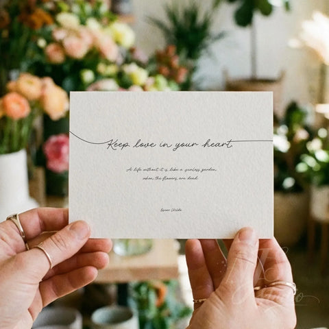 Hands holding a small printed card with Oscar Wilde's love quote, surrounded by soft flowers.