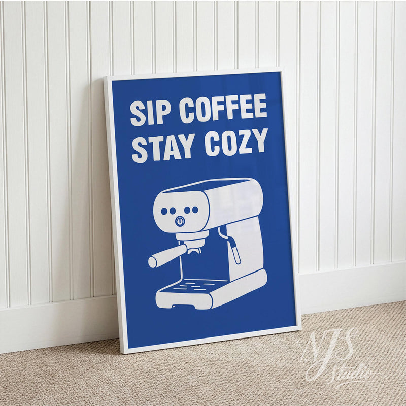Poster with 'Sip Coffee Stay Cozy' text and an espresso machine illustration on a blue background, leaning against a white wall.