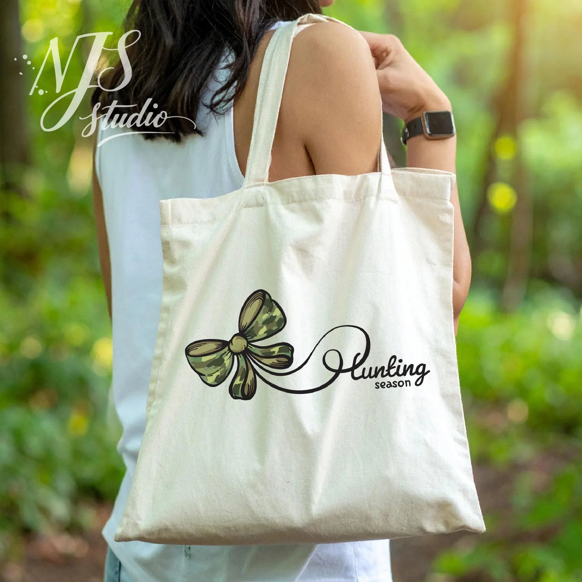 Canvas tote bag printed with a green camouflage bow and “Hunting Season” script, carried outdoors in a natural woodland environment.