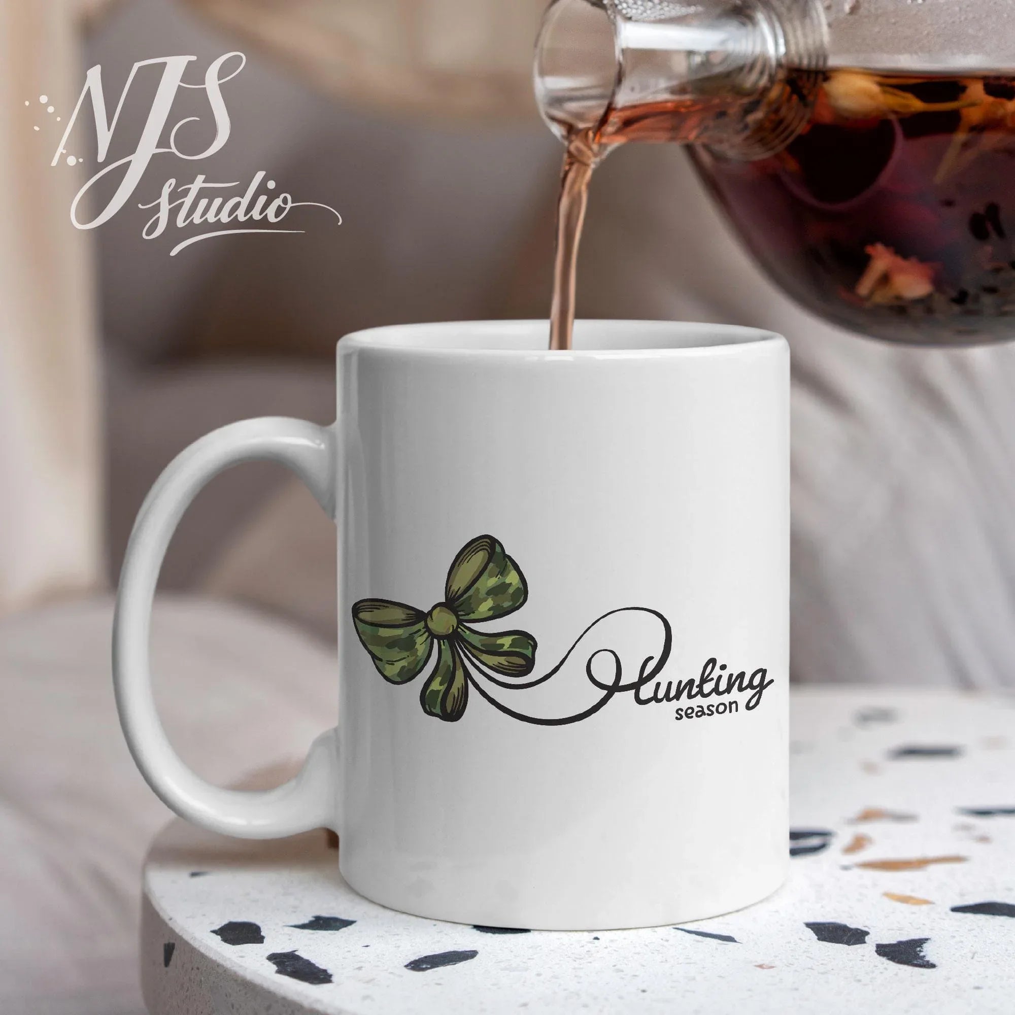 White ceramic mug featuring a green camo bow and “Hunting Season” script as hot tea is being poured in a cozy indoor setting.