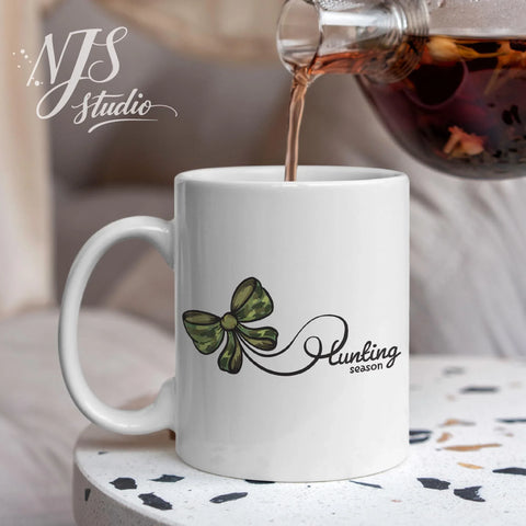White ceramic mug featuring a green camo bow and “Hunting Season” script as hot tea is being poured in a cozy indoor setting.