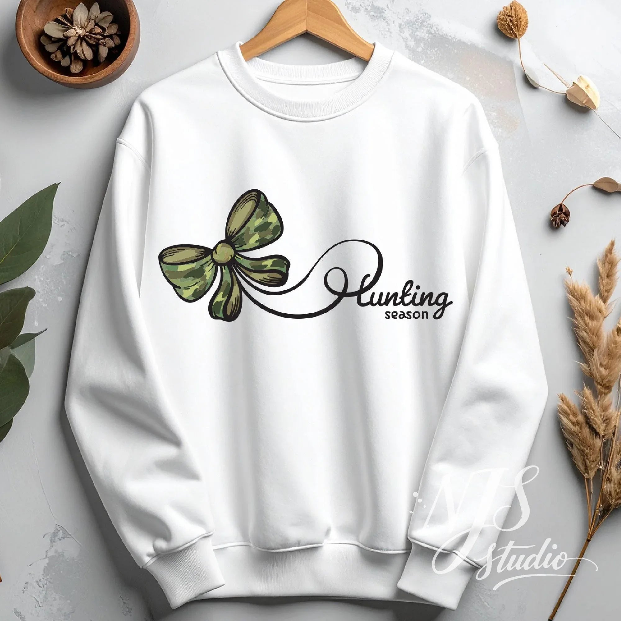 White sweatshirt displayed on a hanger with a green camo bow and flowing hunting season script, styled as a minimal flat lay.