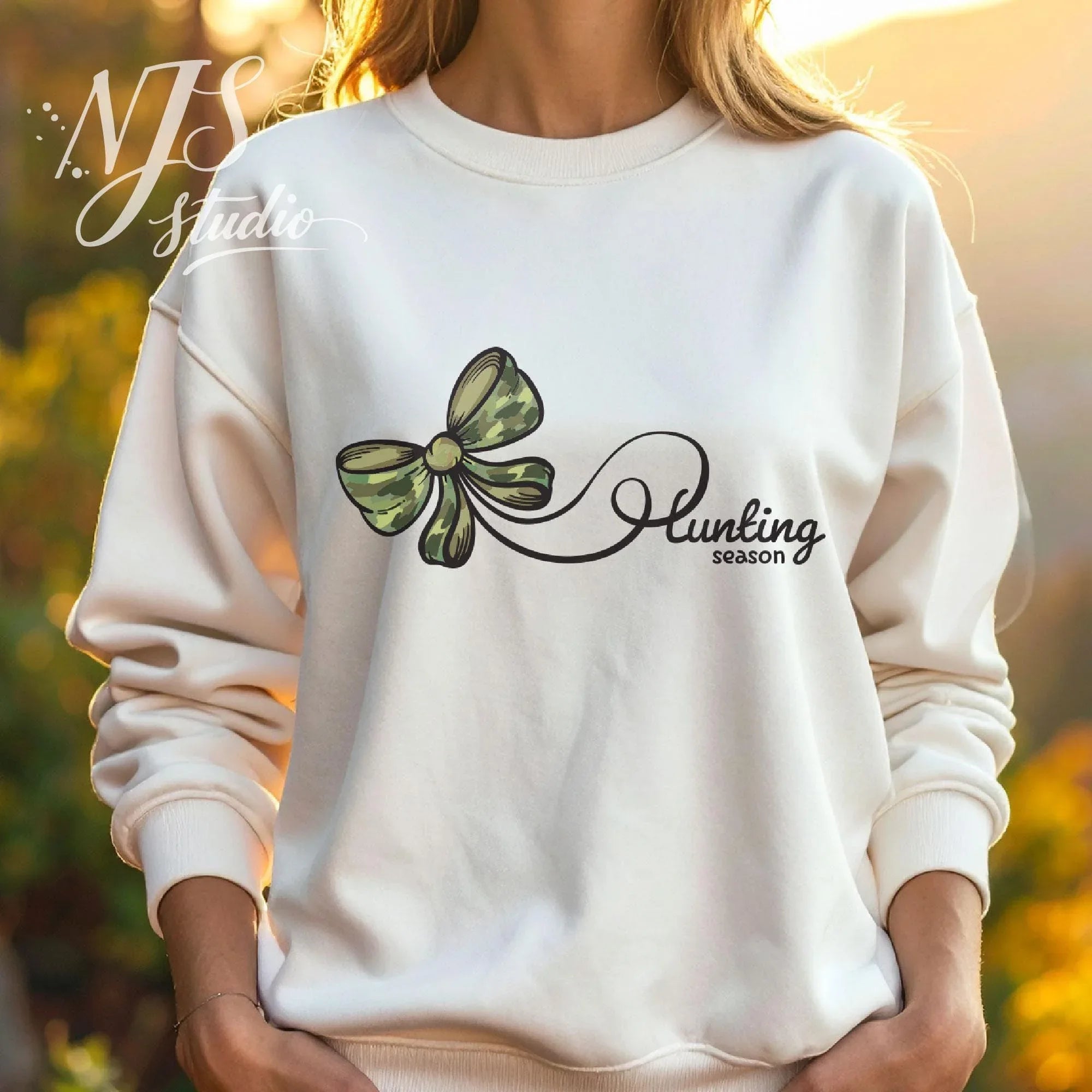 Woman wearing a white sweatshirt with a green camo bow and flowing “Hunting Season” script, styled outdoors in a warm autumn setting.