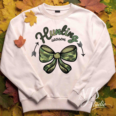 Flat lay of a light-colored sweatshirt with green camo bow and hunting season artwork, styled with autumn leaves.