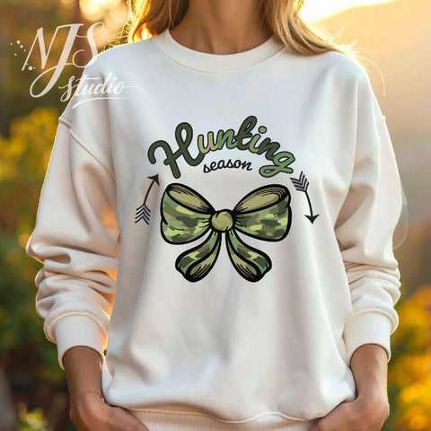 Woman wearing a white sweatshirt featuring a green camo bow and “Hunting Season” design, styled outdoors with a cozy fall aesthetic.