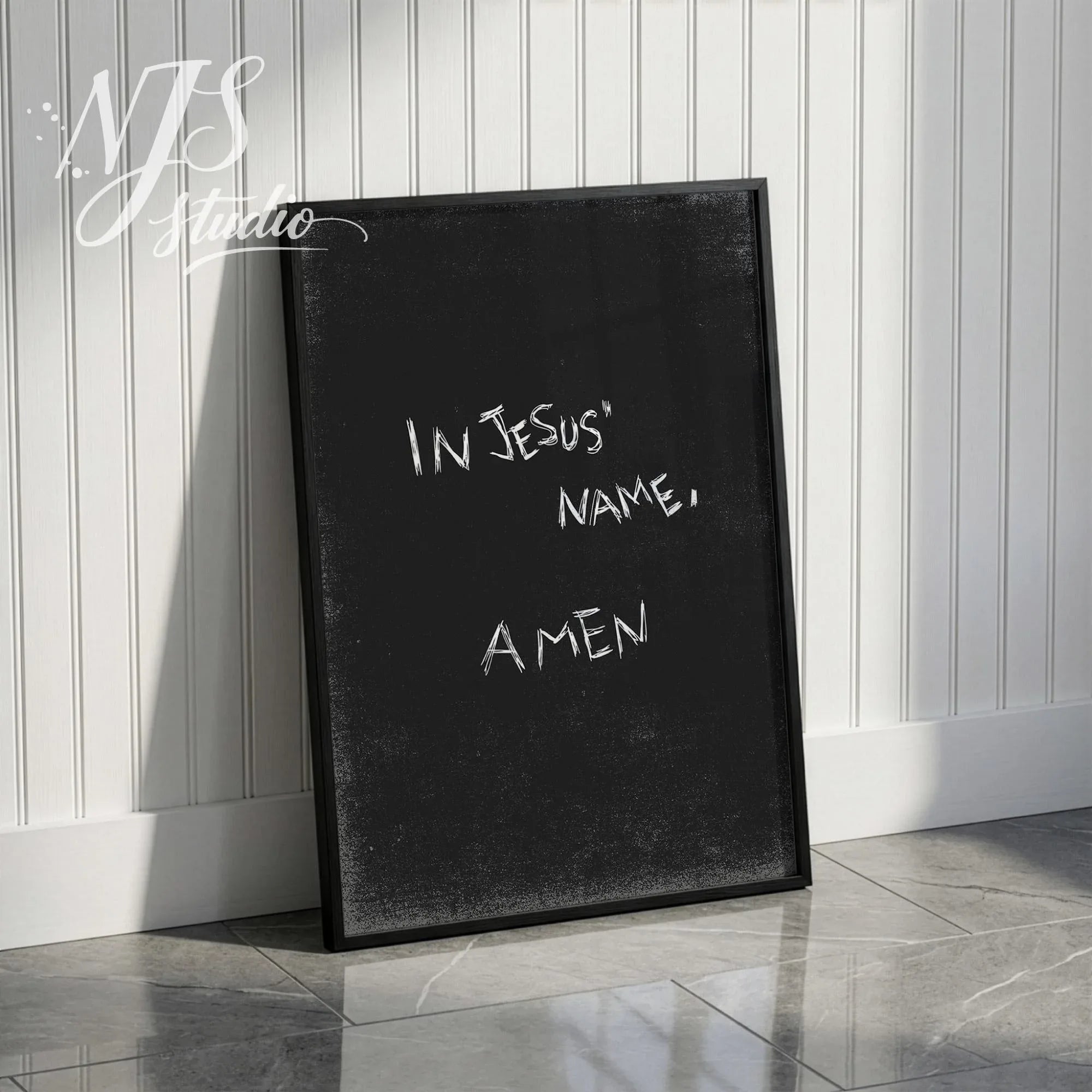 Black framed modern prayer print with text In Jesus Name Amen leaning against a white wainscoting wall.