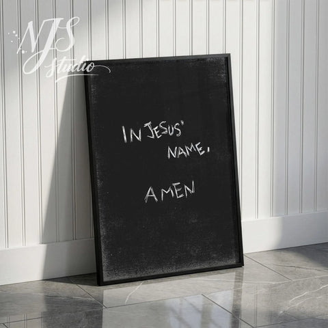Black framed modern prayer print with text In Jesus Name Amen leaning against a white wainscoting wall.