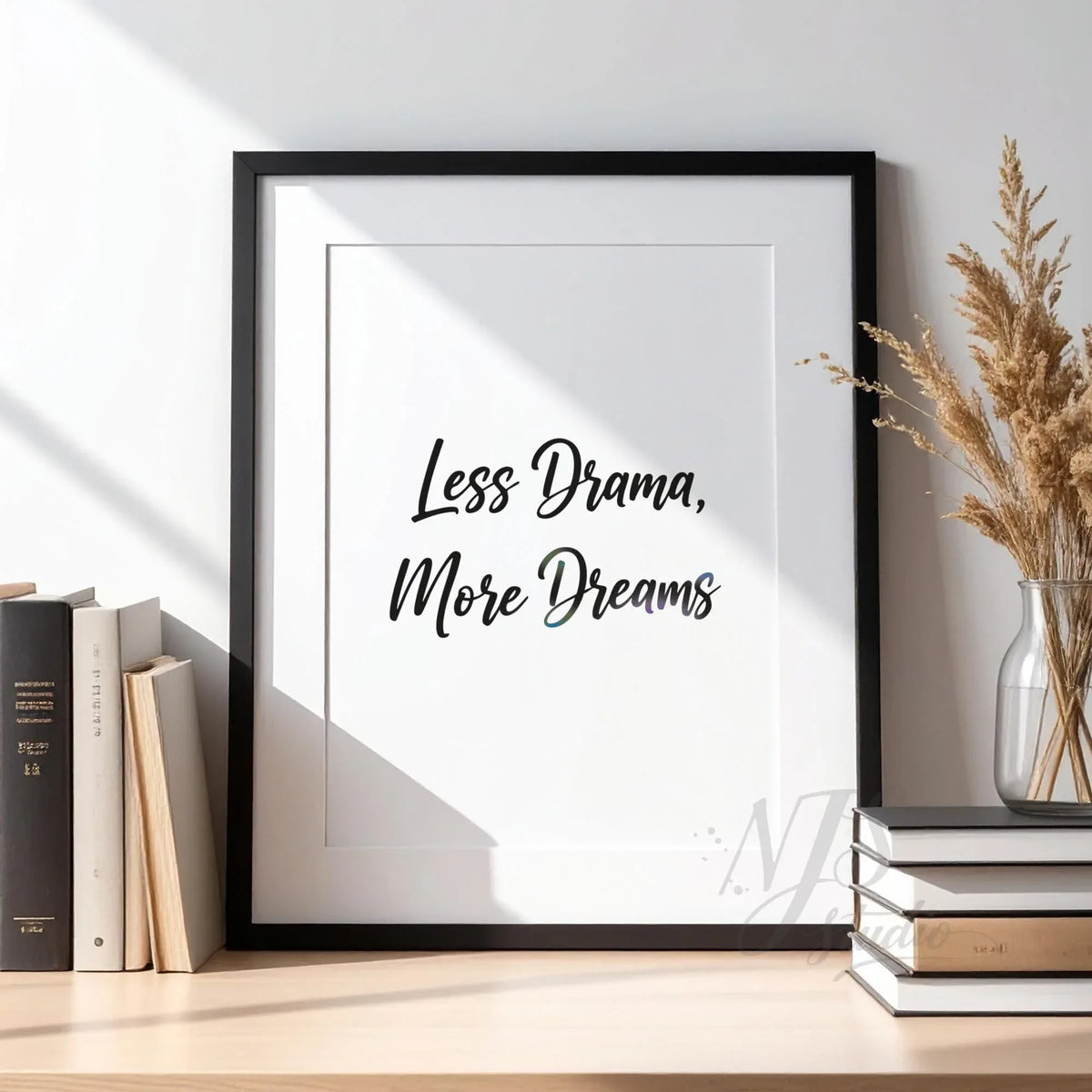 Black framed motivational print standing on a wooden desk with books and dried plants, reading Less Drama More Dreams.