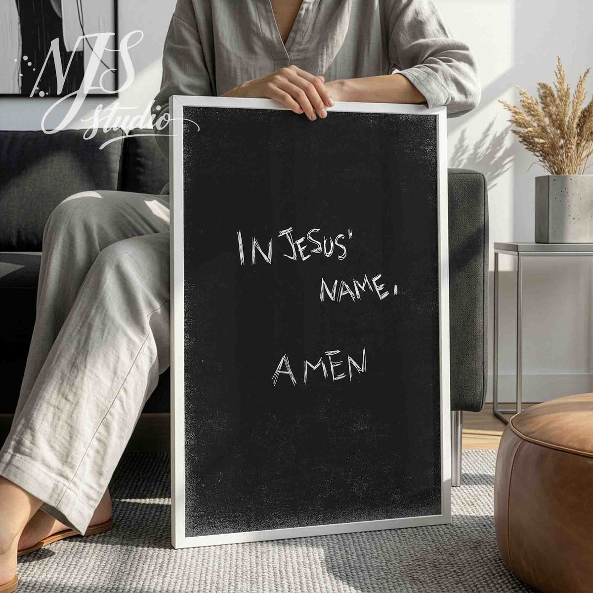 Person in grey outfit sitting on a sofa holding a large white framed black Christian wall art print.