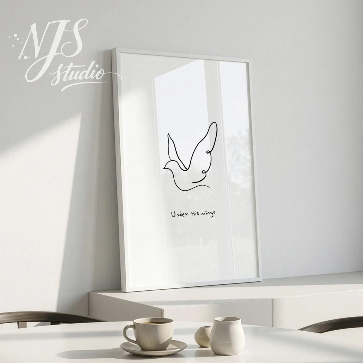 White framed minimalist line art poster of a dove with text Under His Wings leaning on a white sideboard.