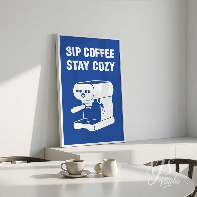 Framed poster with 'Sip Coffee Stay Cozy' text and an espresso machine illustration on a white wall.