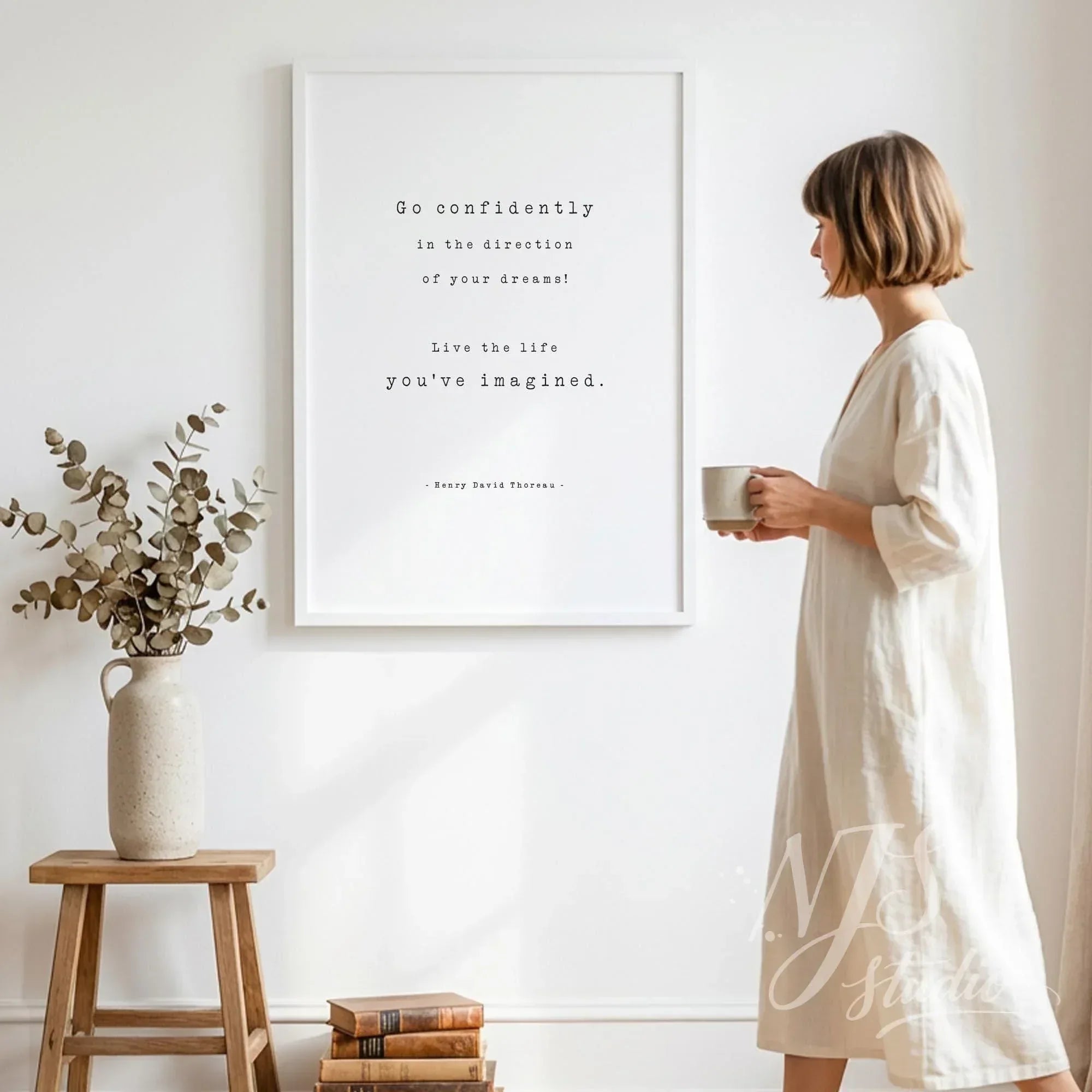 Woman in linen dress holding a coffee mug standing next to a large white framed Go Confidently quote poster in a minimalist home.