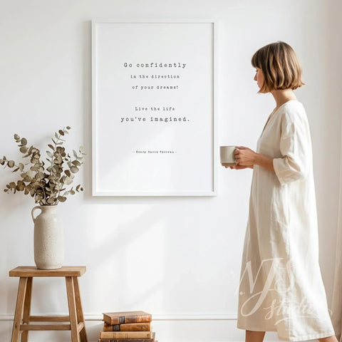 Woman in linen dress holding a coffee mug standing next to a large white framed Go Confidently quote poster in a minimalist home.