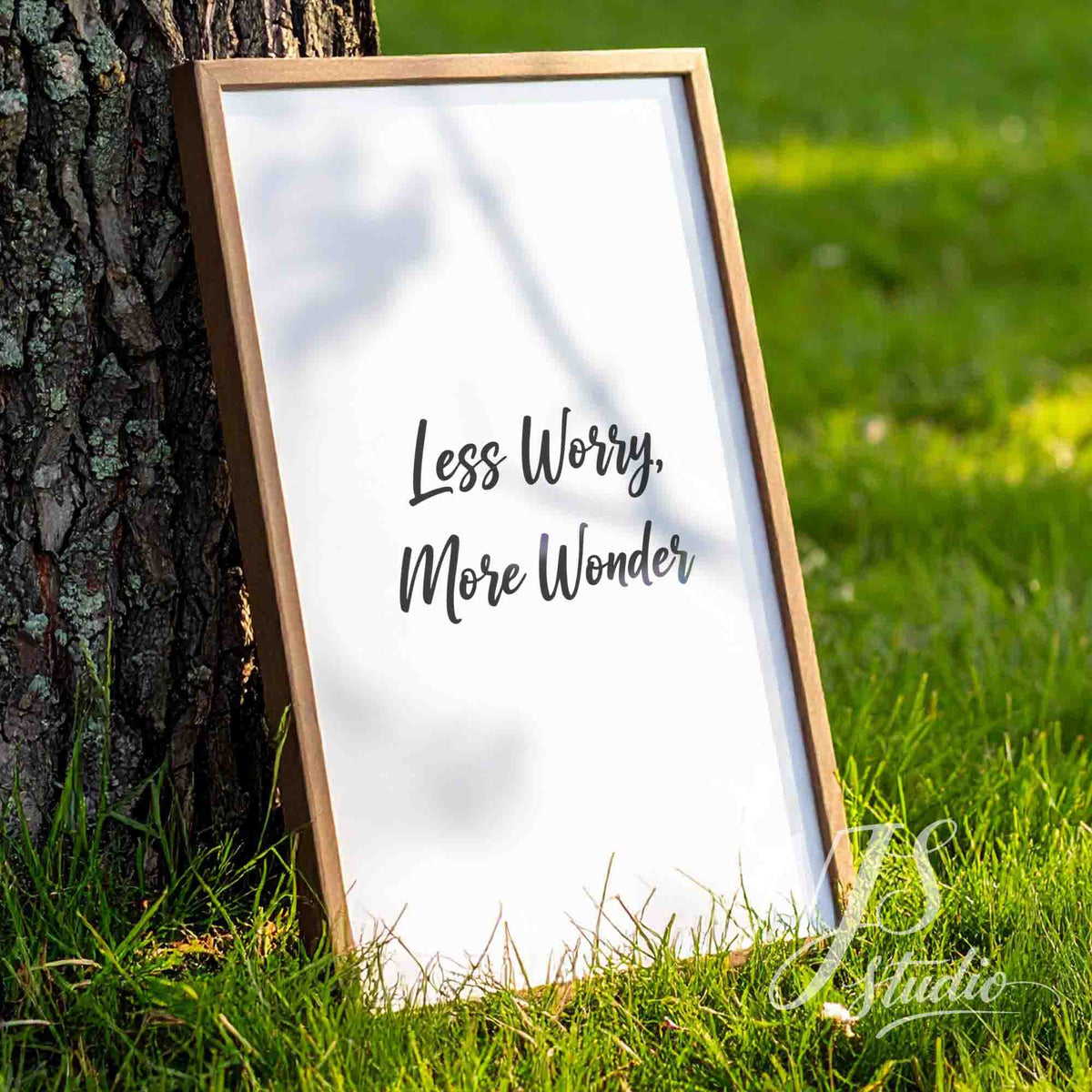 Wooden framed poster leaning against a tree trunk in a garden, sun dappled light, Less Worry More Wonder.