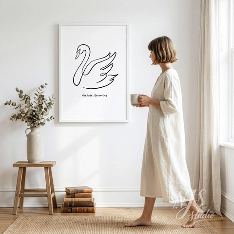 Woman in light dress holding a mug while looking at framed swan line art print hanging on a bright bedroom wall.