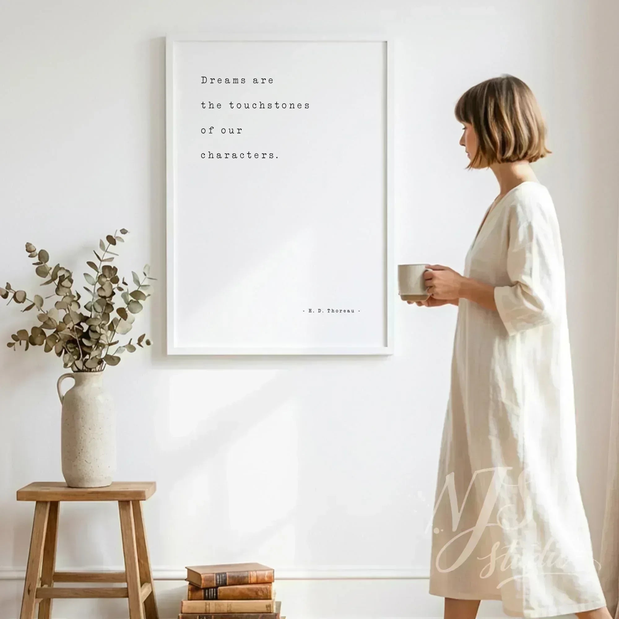 Woman standing in a white dress holding coffee next to a large framed Thoreau quote print.
