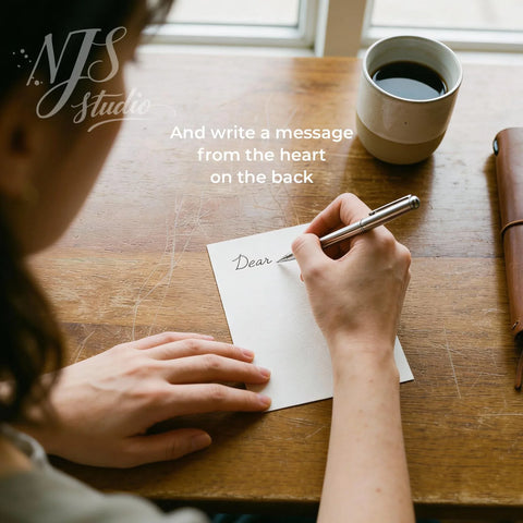 Person writing personal message on card at wooden table, coffee cup, NJS studio branding